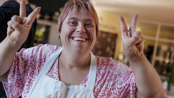 Eine Frau mit Kochschürze lächelt in die Kamera und zeigt mit ihren Fingern das Peace Zeichen