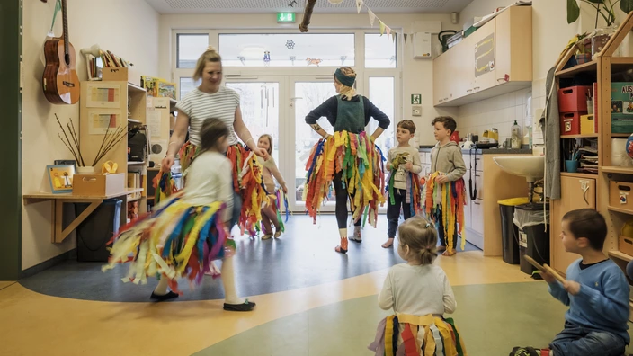 Pädagoginnen und Kinder in einem Kindergarten tanzen in Röcken aus bunten Bändern